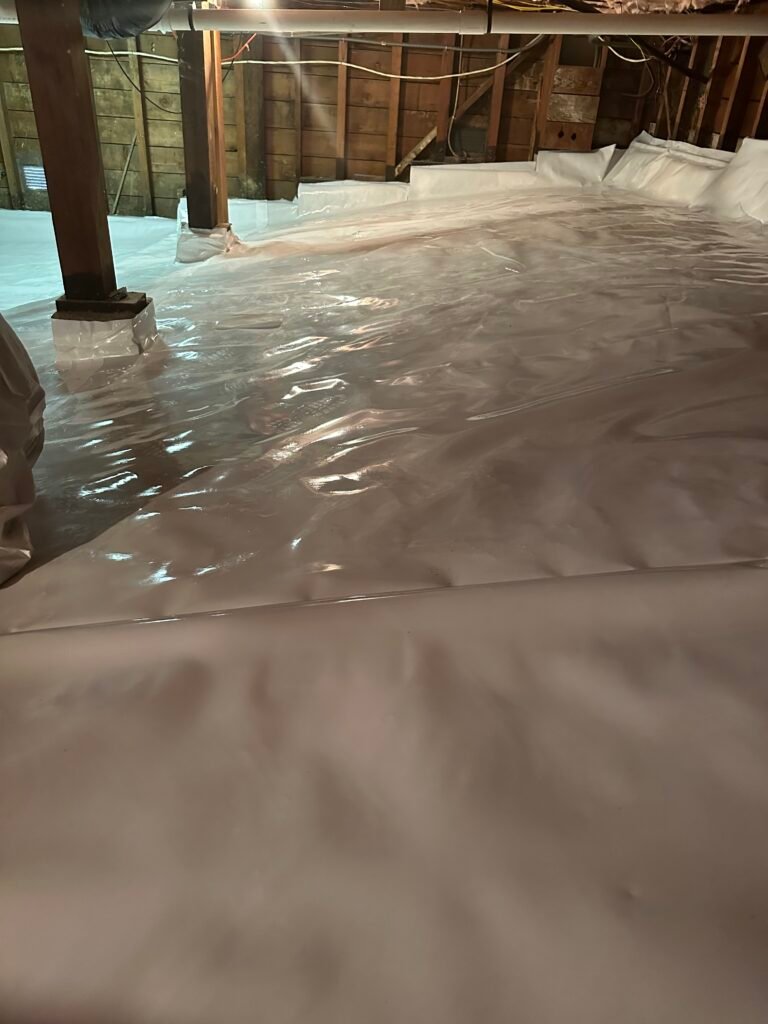 Crawl space with white vapor barrier insulation, wooden beams, and exposed wiring for moisture control and energy efficiency.